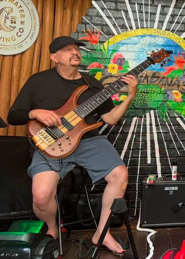 Russ Rodgers seated with his six-string bass in front of the Guanabanas mural